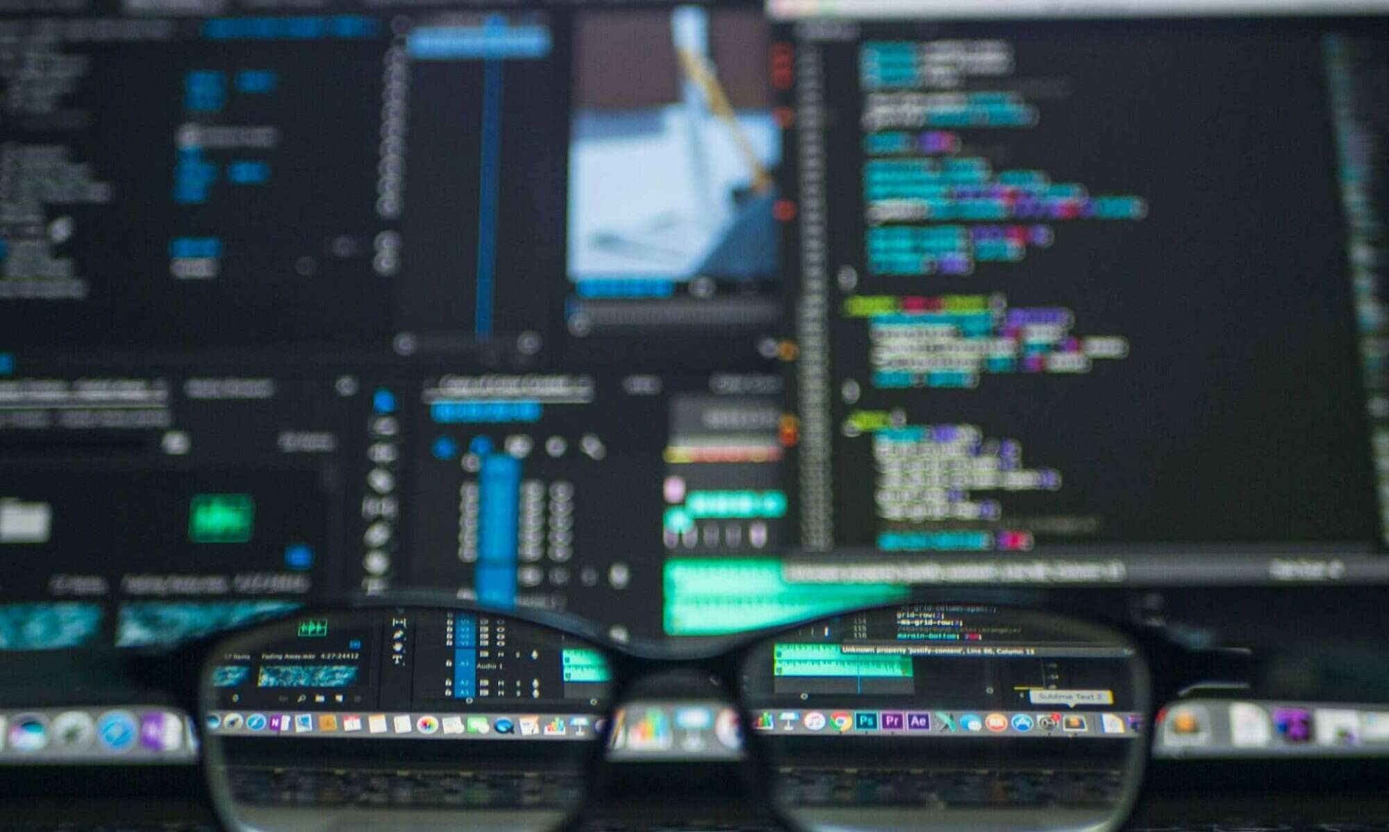 Colorful coding screens displayed on dual monitors with glasses in forefront, highlighting AI office developer activities.